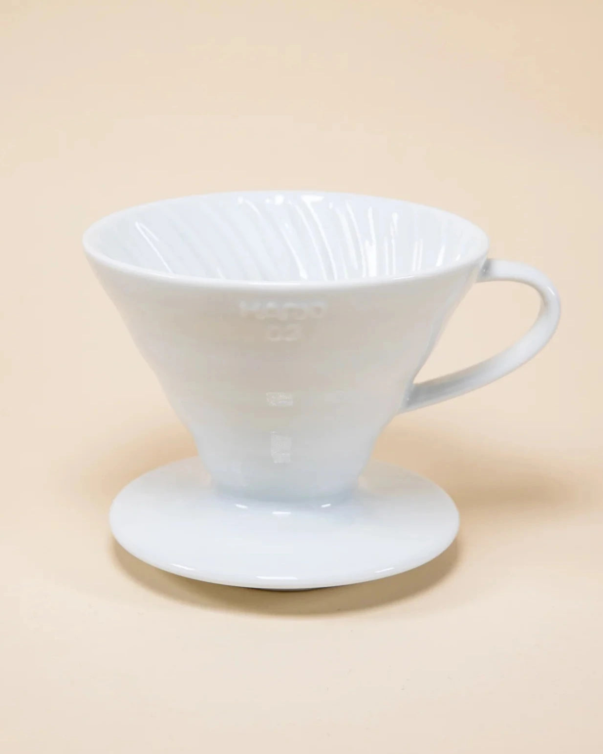 A white Hario V60 ceramic coffee dripper with a paper filter, placed on a matching white stand against a beige background.
