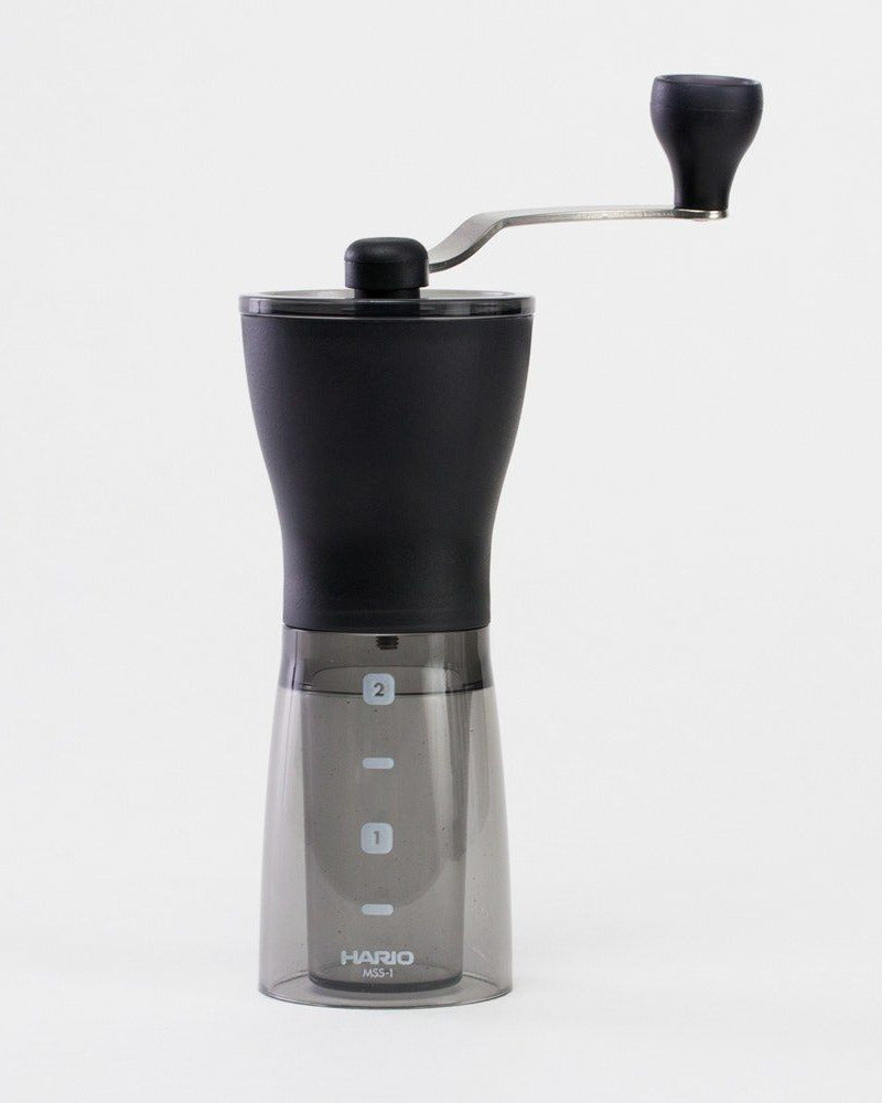 A black Hario ceramic coffee mill with a clear see-through bottle and a knob for adjusting the grind, placed on a white background.